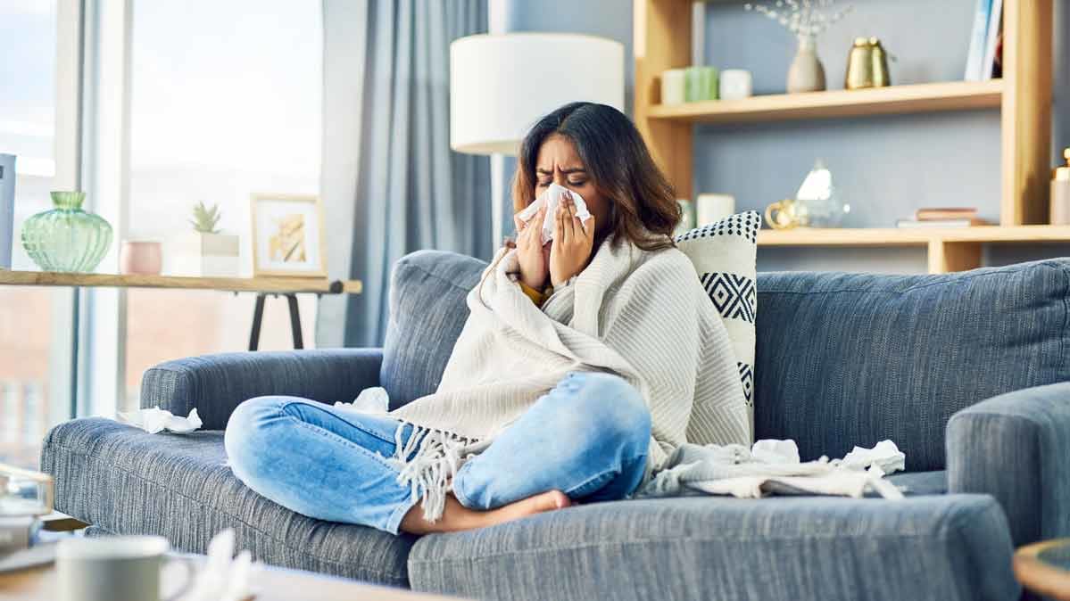 A woman sits cross-legged on a couch, under a blanket, blowing her nose