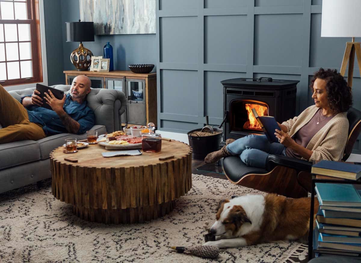 couple reading on couch and chair in living room with Harman Stove Absolute63 pellet stove, dog chewing on toy on carpet