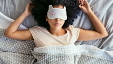 Woman wearing sleep mask covered in blankets in bed.