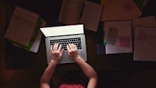 Person using a laptop at night with lots of paperwork on the desk surrounding them.