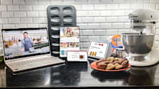 A laptop, tablet, cell phone and smart speaker seen on a kitchen counter with baking accessories.