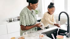 Child washing hands at kitchen sink with woman standing next to her.