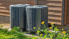 Two heat pumps sit outside a building to the side of green grass