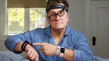 person sitting on edge of edge of bed wearing 6 sleep trackers and pointing to sleep tracker