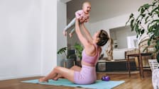 A woman in exercise clothes, sitting atop an exercise mat and next to a pair of dumbbells, holds her baby in the air.