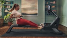 Person using Peloton Rower in living room with built-in bookcases and plants in the background