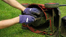 detail of person wearing gloves scraping out grass clippings from underneath a ride behind lawn mower