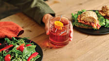 Hand holding a cocktail glass with an orange peel, and plates with salad and sandwiches on a wooden table.