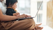 Toddler sitting in their parents lap looking at laptop.