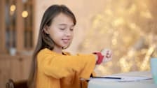 child with long brown hair wearing yellow shirt, sitting at desk and looking at their red smartwatch