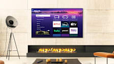 A Roku-branded TV in a living room setting displaying the home screen