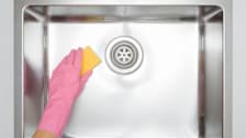 Hand in pink rubber glove cleaning stainless kitchen sink with sponge.