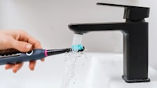 close up of hand holding electric toothbrush under bathroom faucet