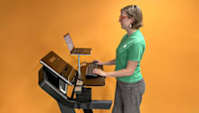 person wearing green CR branded t-shirt using laptop while standing on treadmill with RiseEight treadmill desk attachment in place