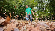 person wearing green Consumer Reports shirt while using leaf blower, leaves in foreground and trees in background