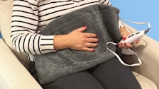 close up of Pure Heating Pad on person's lap, person sitting in beige chair holding heading pad control in one hand