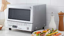 Panasonic FlashXpress NB-G110P microwave on white countertop with white subway tile backsplash, plate of roasted vegetables with cheese to right of microwave