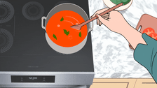 Hand stirring tomato sauce with basil leaves in a pot on an induction cooktop.
