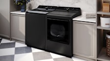 An LG WT8480CL top-loading washer and matching LG dryer in a laundry room with gray cabinets and a black-and-white tile floor.