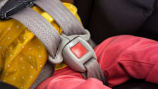 Close-up of a five-point harness in a child car seat, with the child wearing coral-colored pants and a yellow shirt with white polka dots.