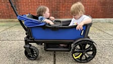 two toddlers in blue Veer baby stroller wagon on concrete sidewalk with brick wall behind them