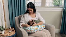 A mother sitting while breastfeeding her newborn baby with the New Boppy Nursing Pillow in Colorful Forest Animals print.