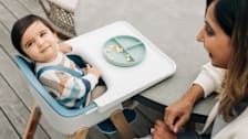 baby in striped sweater sitting in Uppababy Ciro high chair at round table, with adult smiling at them in corner of frame