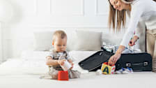 Mother packing a suitcase on a bed while infant plays next to her.