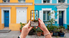 A person's hands holding a smartphone, taking a photo of two colorful building facades.