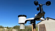 Ambient Weather weather station on wooden post with blue sky, desert landscape, and building in background