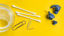 blue earbuds with three cotton swaps, two paperclips, and small bowl of soapy water on yellow background