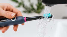 close up of dark blue and black electric toothbrush under faucet with stream of water hitting toothbrush head