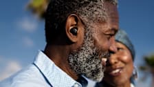 side view of person smiling while wearing black Sony hearing aid, second smiling person blurred in background