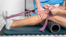 Person using a resistance belt and foam roller on fitness mat while physical therapist offers guidance