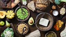 overhead view of wooden bowls and boards with beans, nuts, tofu, whole grain bread, squash, broccoli, cauliflower, avocado, and dried fruit