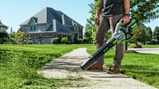 a man using a Ego 56V battery handheld leaf blower on a residential sidewalk to blow grass clippings.