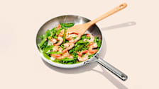 Oxo Tri-Ply Stainless Mira Series frying pan with wooden spoon, shrimp, peas, and sugar snap peas on light pink background