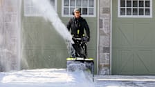 A woman using a Ryobi RY408150 Two-Stage Battery Snow Blower to clear snow off a residential driveway.