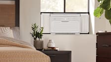 Frigidaire Gallery GHWQ103WC1 window air conditioner, corner of bed on left side and nightstand under the air conditioner with ZZ plant, vase, and two books