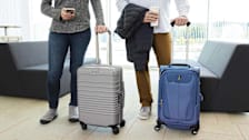 low view of two people with carry-on luggage, holding coffee container and phone, with couches and windows behind them