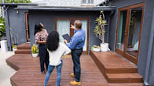 couple looking at home with home inspector holding tablet, pointing to roof of house while standing on wooden deck