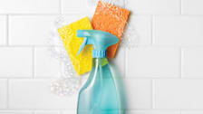 Blue spray bottle against white subway tile with a yellow and orange sponge and bubbles.
