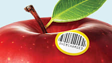 A close-up of a red apple that features a barcode sticker that conspicuously displays "OVERCHARGED"