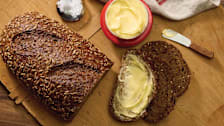 Seeded rye bread loaf and buttered slices with container of butter and salt on a wooden board.
