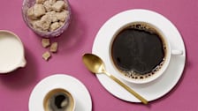 A top-down view of a cup of coffee, an espresso, a creamer pitcher, and a jar of brown sugar cubes on a purple surface.