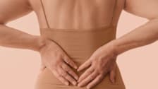 A person's hands are placed on their lower back while wearing a neutral-toned top.