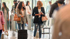 Two women with naturally curly hair walking with their luggage and carry on bags through an airport.