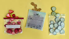 An Amazon Basics bag holding strawberries, a GreeneWrap bag with pretzels, and a Glad Flex’N Seal bag containing cucumber slices on a yellow background.