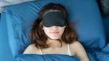 person sleeping on back while wearing sleep mask, blue pillow cover and sheets on bed