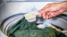 Hand holding a scoop with powered laundry detergent over an open washing machine.
